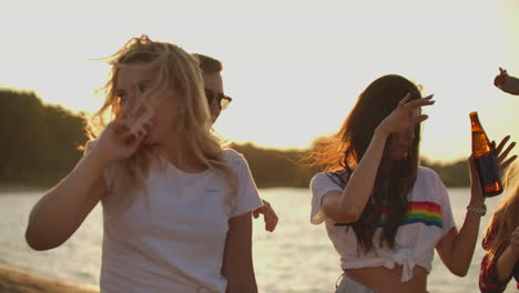 Three-female-student-are-dancing-in-short-t-shirts-with-beer-on-the-beach-party-at-sunset.-Their-long-hair-is-flying-on-the-wind.-This-is-hot-and-carefree-summer-evening.